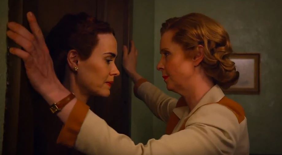 Mildred stands looking down with her back against the wall as Gwendolyn traps her in with her arms on either side of the doorframe.