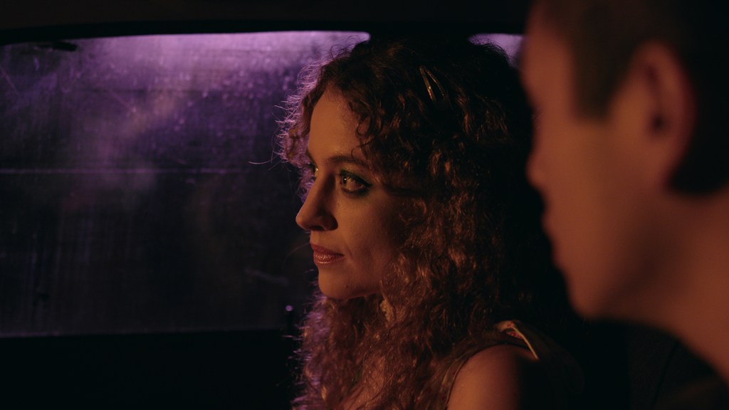 A woman and man sit in a car. The window is foggy, and the characters are lit by reddish purple lighting.