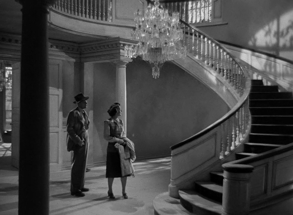 A black and white photo of a man and woman in semi-formal 1940s attire standing in the entry of a mansion with a large curved staircase and chandelier