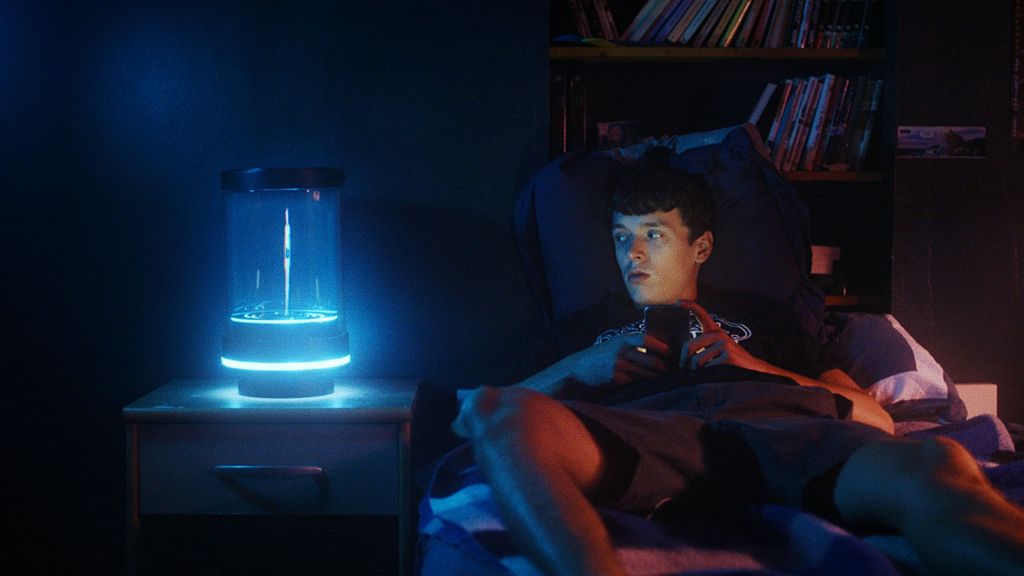 A teenage boy sits on a bed in a dark room, looking to his right at a tube containing an AI girl.
