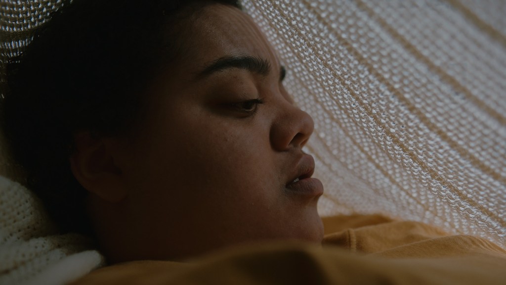 Still from Our Sister: Tasha, a nonverbal autistic teen is wearing a yellow jumped and is lying under a crocheted white blanket. she looks upset. 