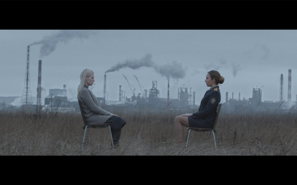 Still from Sulphur: Two women are sitting in a field. The woman to the left is blonde in a grey cardigan while the brown-haired women on the right is a policewomen in uniform. In the background are factories with smoke going into the sky above. 