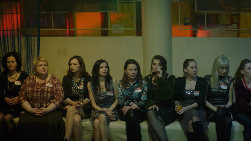 A row of different women sat on a bench inside a dark community centre, all wearing name badges. From 'Anna'