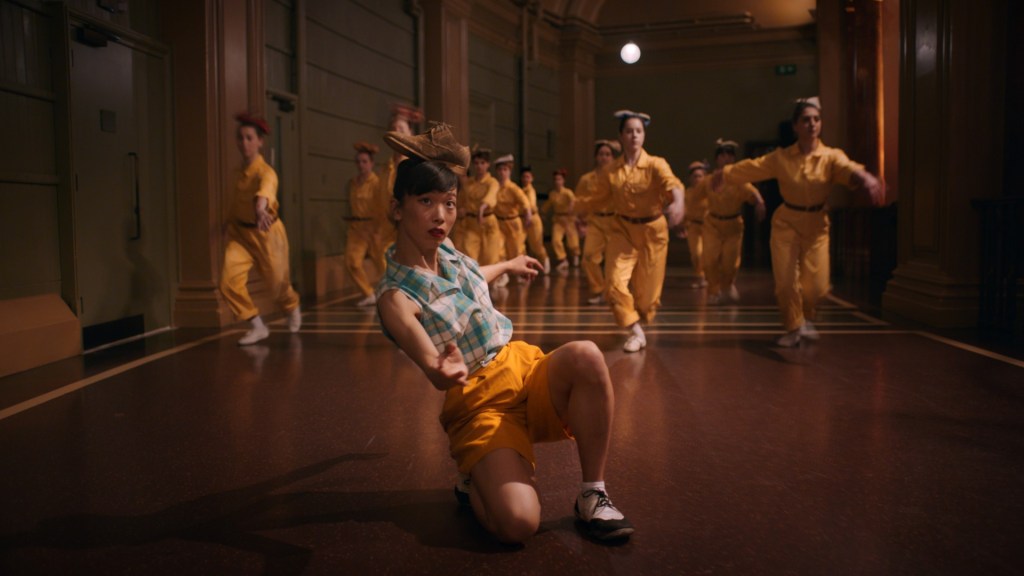 A woman in a checkered blue top and yellow shorts kneels on the ground balancing a brown shoe on her head. In the background is a chorus of dancers in yellow work uniforms. From 'Cinderella Games'.