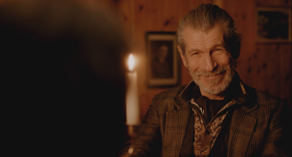 An elderly man- wearing a formal, floral shirt and a tweed jacket- smiles to someone off screen at a candlelit dinner. From 'Brothers Again'