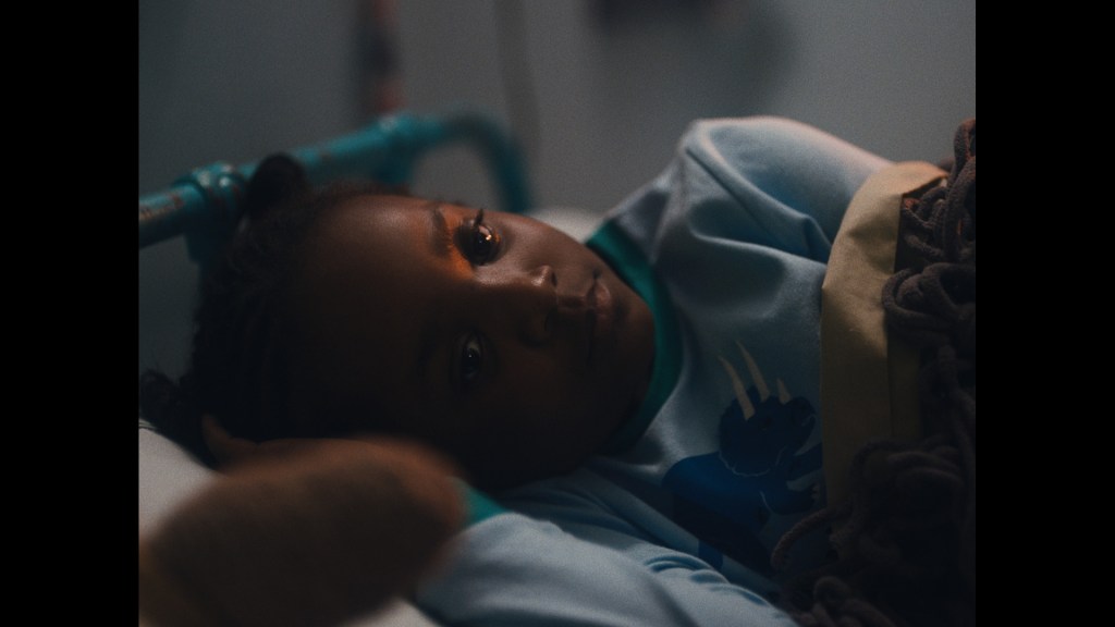Still from Dawn in the Dark: Dawn, a young child is tucked into bed. Her dark skin is lit by her nightlight, covers wrapping her up.