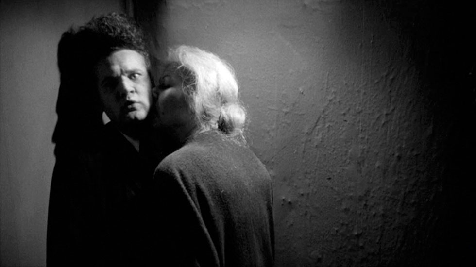 Black and white; A white man with a curly flat top haircut is kissed on the neck by a white woman with light blonde hair