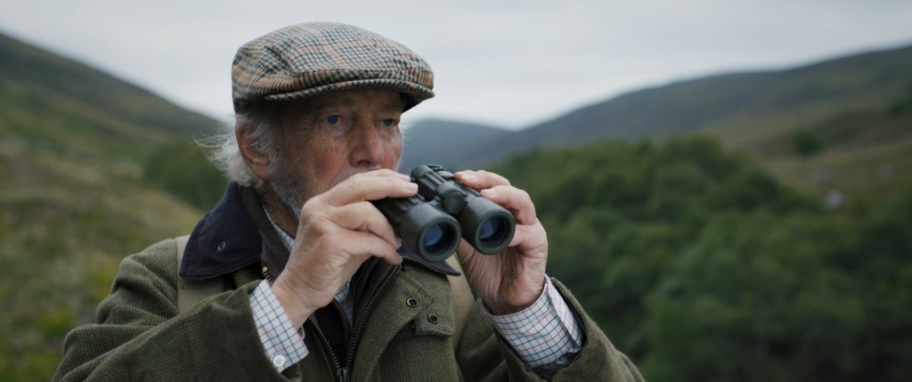 An elderly man in a typical farmers jacket and flatcap, lowers his binoculars and gazes at something off screen. He is standing in the rolling hills of the Scottish Highlands. From 'Ghillie'