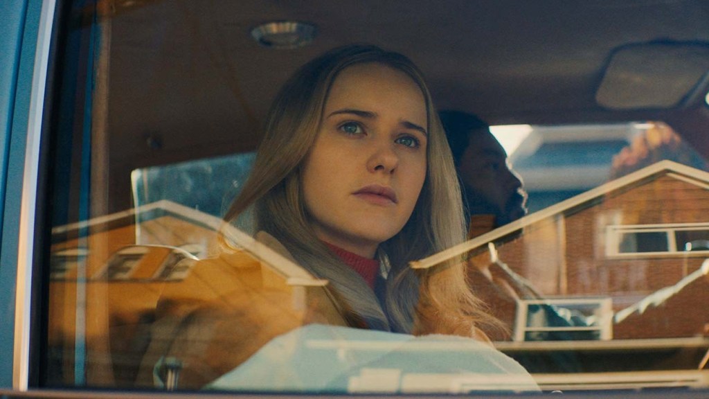 A blonde woman staring out of the car window from the passenger seat. Sitting on her left is a man who drives the car.