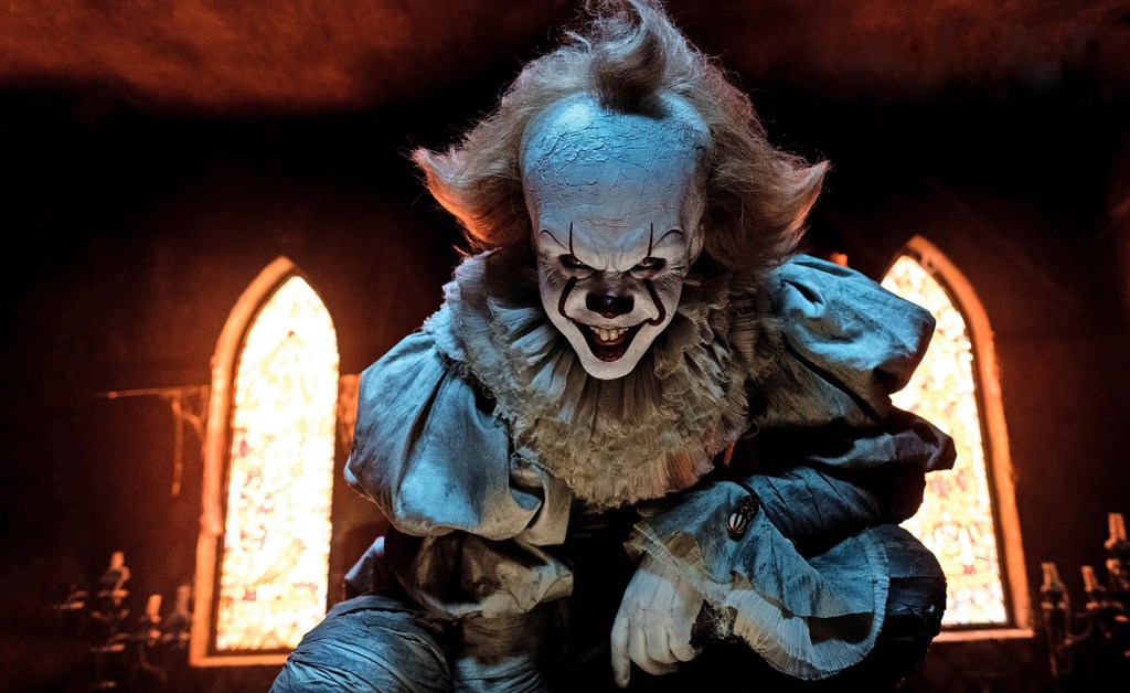 Pennywise the clown smiles creepily in blue lighting with two windows glowing behind him