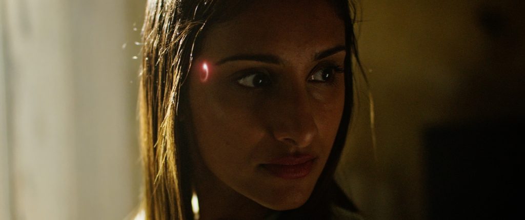 A woman in a dark, nondescript room, turns her head to glance at something off screen. A bright orange circle glows on her right temple. From 'Muse'