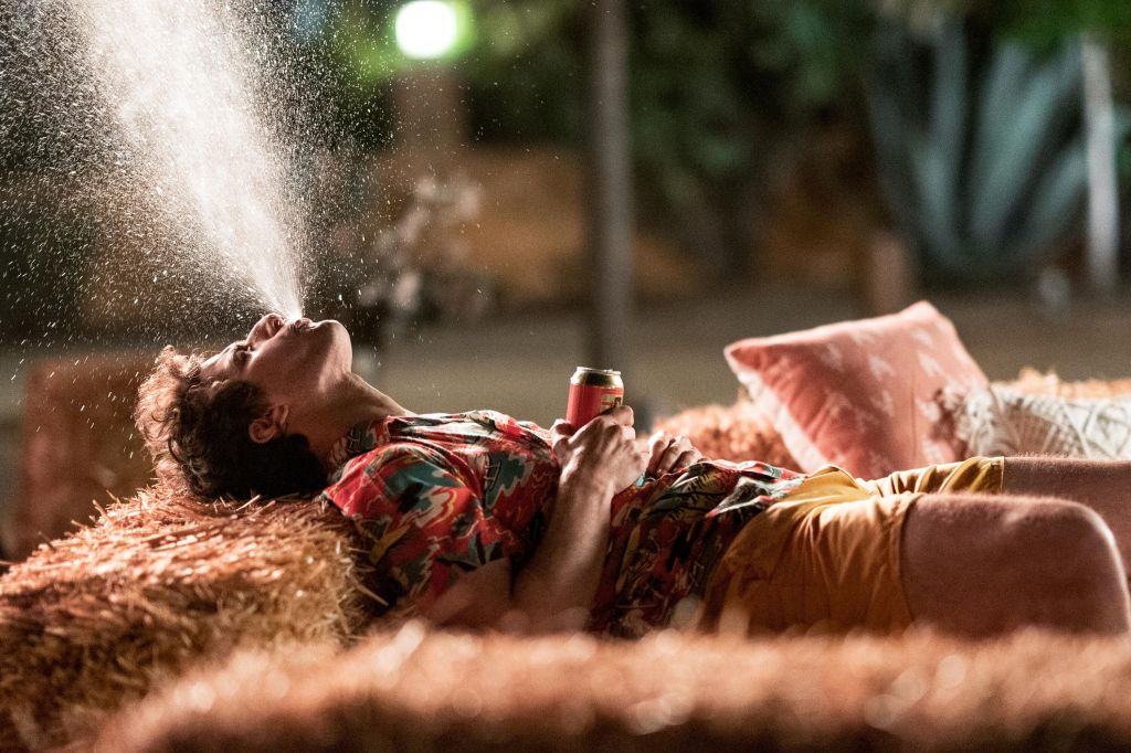 This image is from 'Palm Springs' (2020). Nyles (Andy Samberg) lies back on a hay bale and sprays water into the air.