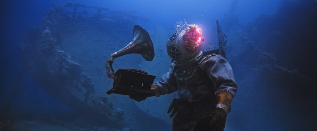 In the dark depths of the sea, in front of a wreckage, a deep-sea diver in a 20th century diving suit holds up a vinyl player. From 'The Coral Guardian'.