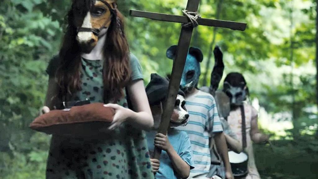 5 children wearing various animal masks walk in a line in the woods. The first holds a pillow, the second a homemade cross, and the last a drum. 
