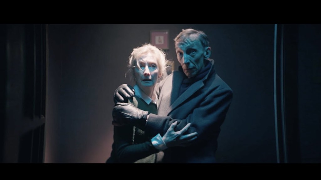 An elderly couple (played by Shelia McCarthy and Julian Richings) hold each other in panic. They're standing in a dimly lit hallway. The woman has dishvelled white hair and the man has thin receeding hair. The man looks calmer than the woman, who is more alarmed by what she is looking at.
