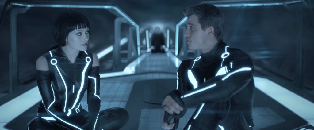 A women and a man, each wearing skin-tight suits laced with strips of blue neon lights, sit on a futuristic space deck whilst looking at each other. In the background out of focus is a figure in black clothing kneeling as if in prayer. From 'Tron: Legacy'