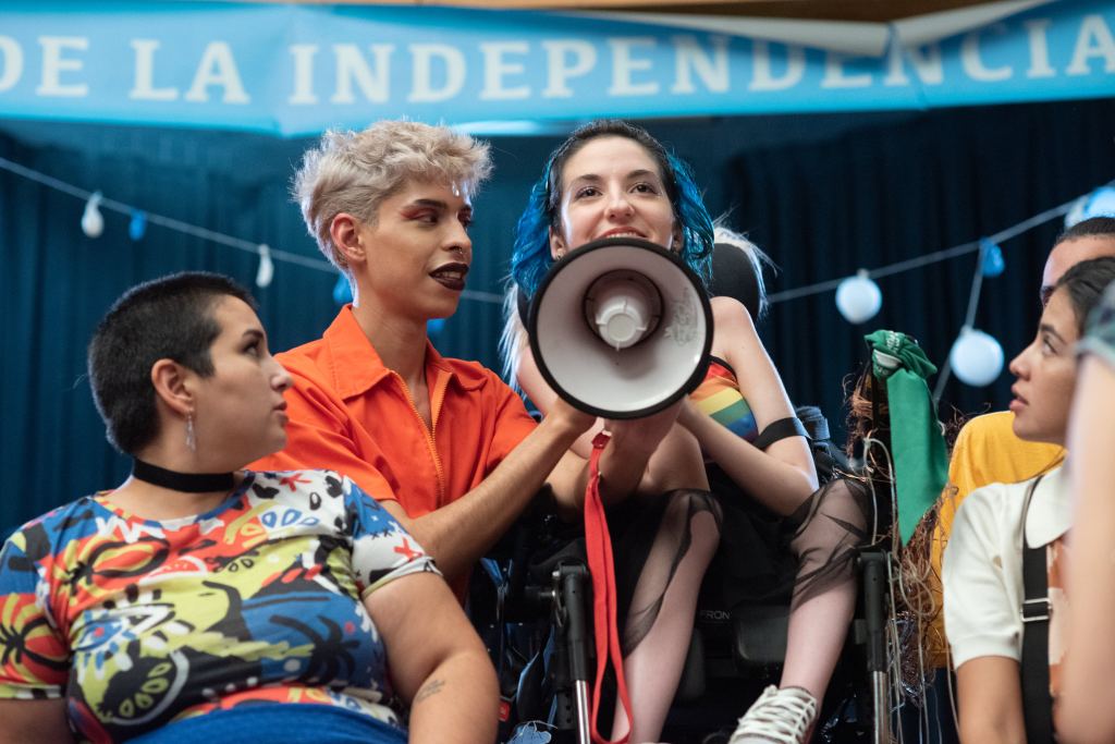A young, female wheelchair user shouts into a megaphone which is held up by her friend.