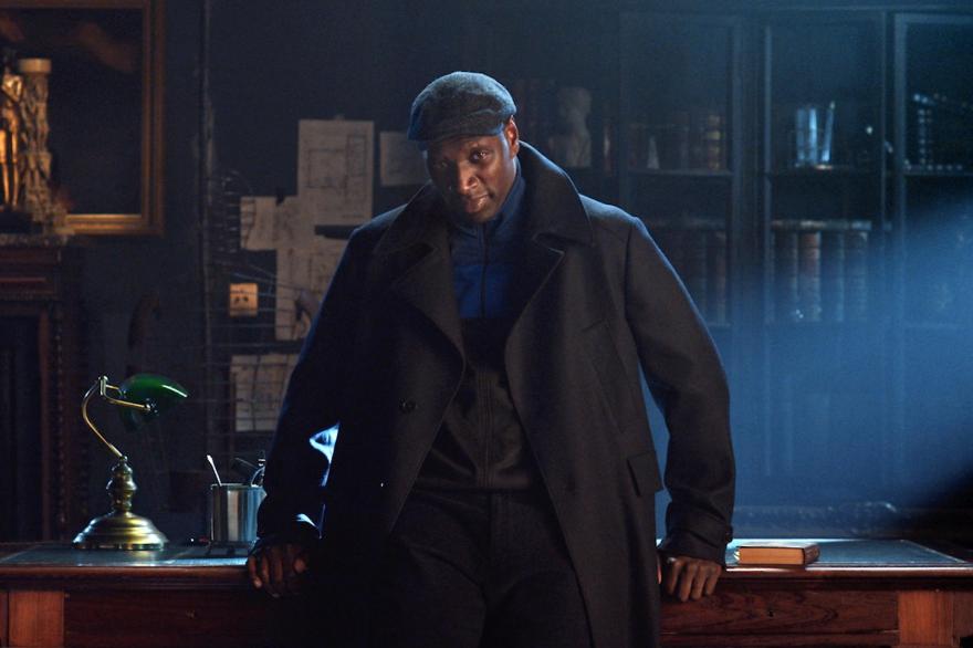 Omar Sy as Assane Diop, dressed in a big black coat and leaning against a desk.