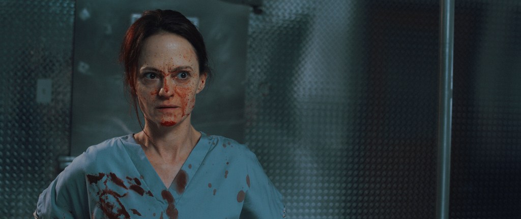 Angela Bettis in horror-comedy 12 Hour Shift. A woman in a light blue nurse uniform looks alarmed and annoyed, her gown and face are covered in blood. The background is a grey, bluish wall of an autopsy room.