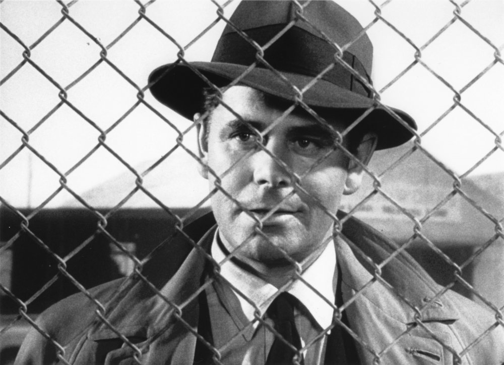 Dave Bannion (Glenn Ford) stands behind metal fencing.