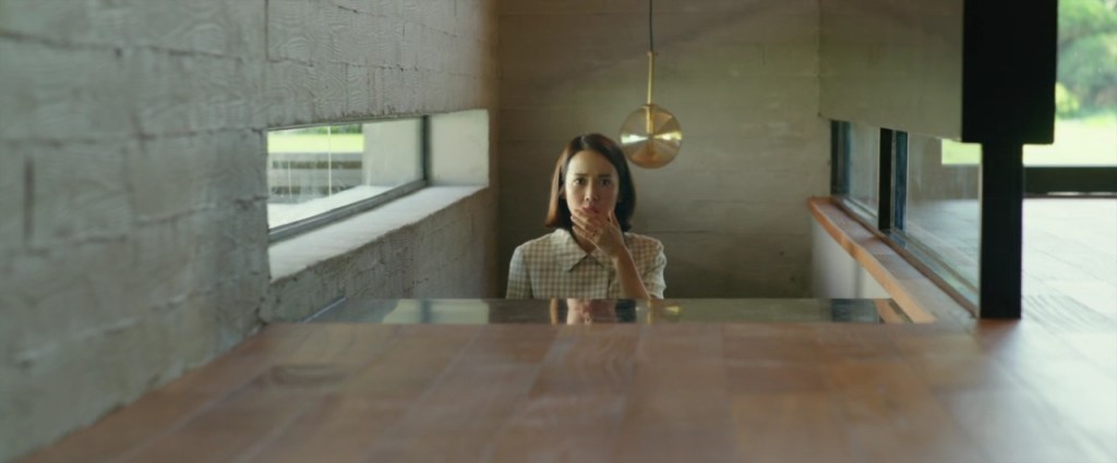 We see the top half of a woman wearing a bright white and yellow top as she makes her way up some stairs in a sleek, modern and minimalistic house with wooden flooring. Her hand is held over her mouth, shocked at something off screen. 