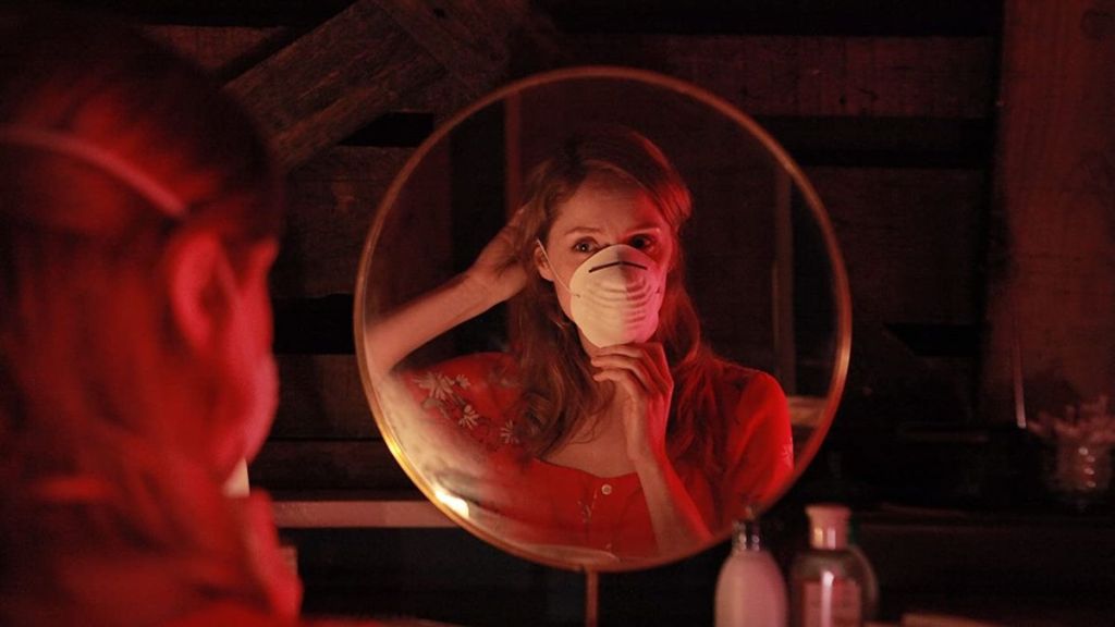 A circular mirror reveals a young woman pulling a respirator mask over her face, bathed in a dim, red light.