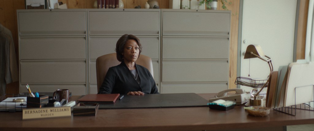 A concerned looking woman sits at a large wooden desk adorned with folders, a phone, stationary and a name plaque saying "Bernadine Williams- Warden"