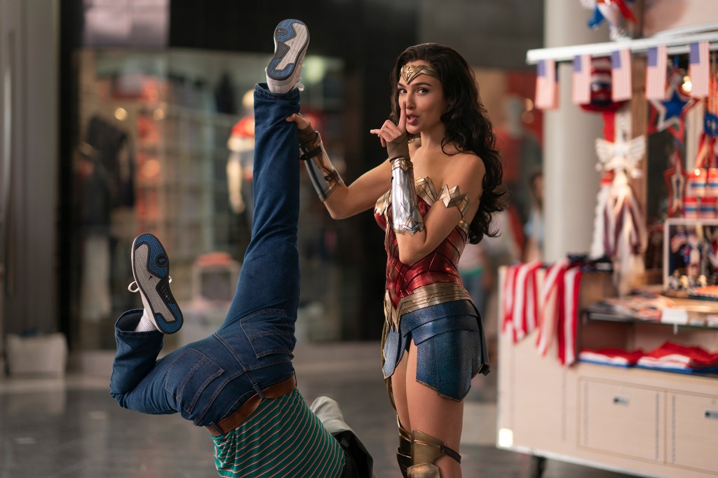 Wonder Woman (Gal Gadot) holds a person up by the legs with one hand, with her finger to her lips in a "shush" gesture.