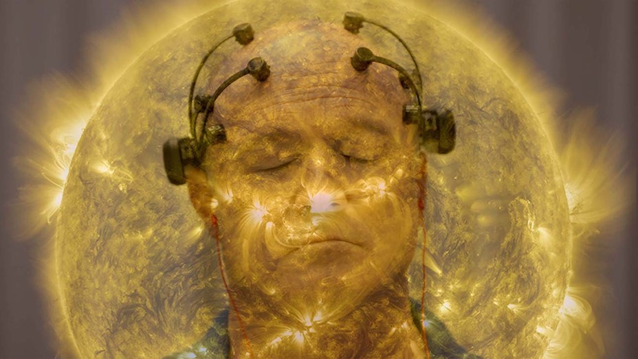 The image from the documentary All Light Everywhere. It shows two separate images layered over each other. The first a yellow hued image of the sun, and the second a man with scientific equipment attached to his head.