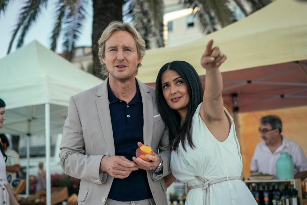 A still from the sci-fi film Bliss. The background is a sunny seaside resort with a blurred palm tree behind a confused man and happy woman. The man has blonde hair and is played by Owen Wilson, dressed in a grey suit jacket with a black shirt. He's holding a half-eaten peach. The woman has long black hair, dressed in a white gown, and is played by Salma Hayek. She's pointing proudly into the distance at something we cannot see.