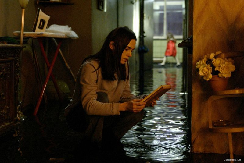 Image from the film 'Dark Water'. A young woman kneels down in a corridor that is completed flooded, holding a book. At the end of the corridor, a little girl wearing a red coat walks past.
