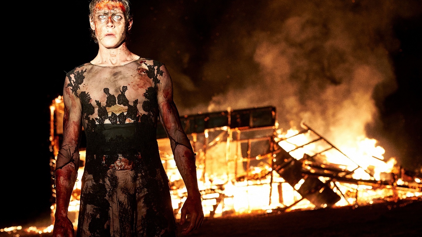 Ned Kelly (George Mackay) stands bloodied in his Rorschach-esque dress. Behind him, a building is burning to the ground.