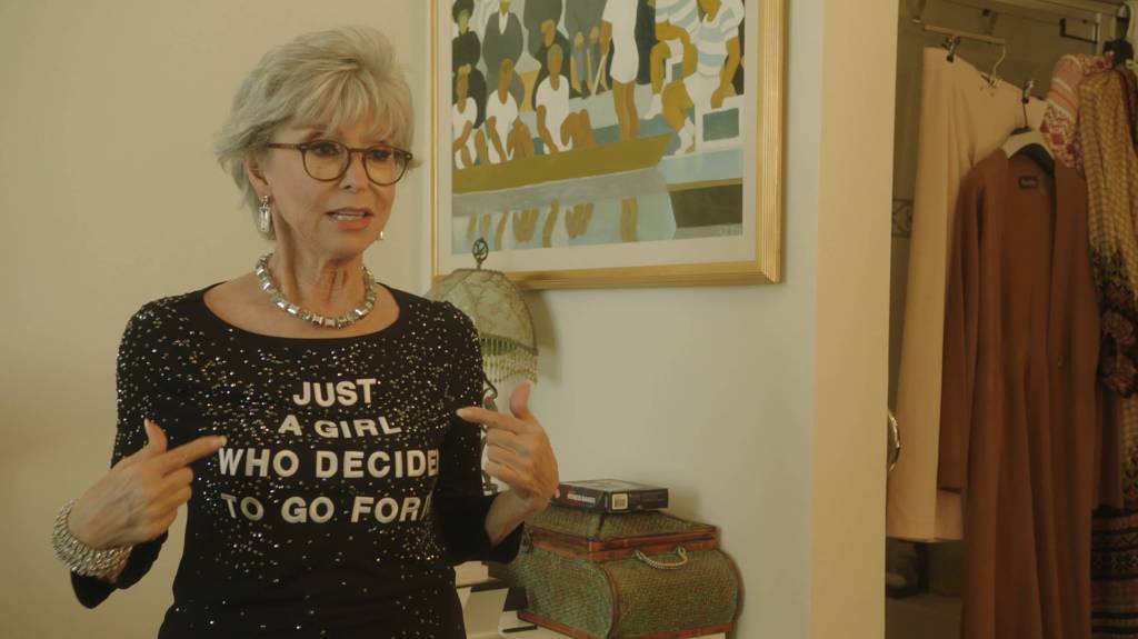 Rita Moreno points to her bedazzled shirt that reads "Just a Girl Who Decided to Go for It"