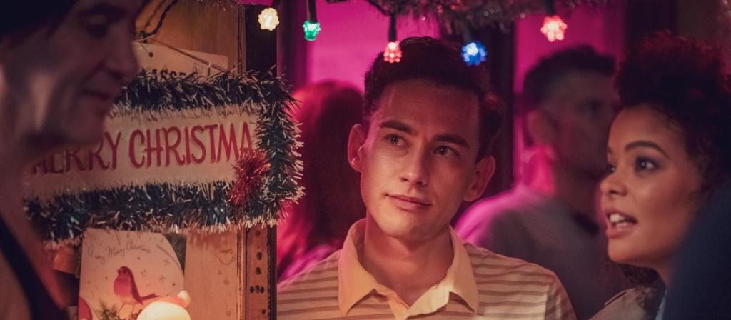 The characters of Richie and Lydia stand at a bar surrounded by brightly coloured lights whilst Richie looks thoughtfully ahead.