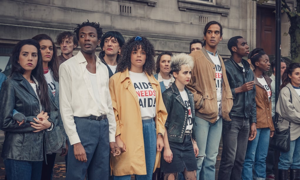 Lydia and Roscoe nervously stand with a group of their friends at a protest wearing 'AIDS need aid' t-shirts.
