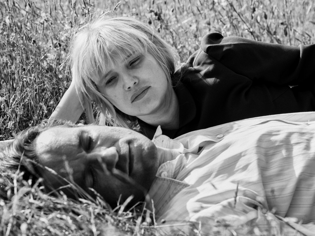 Wiktor and Zula lay in a field. Zula lays on her side and watches him. He is wearing a white shirt with stripes whilst she wears black. The image is in black and white.