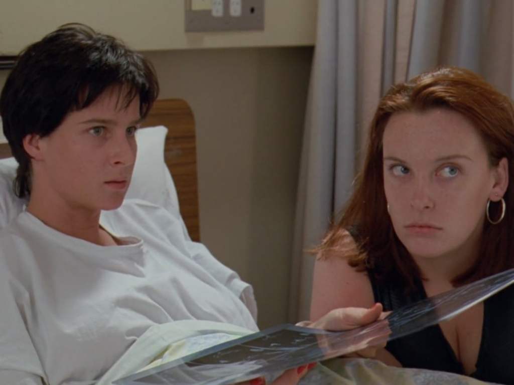 Image from the film Muriel's Wedding. Rhonda, a woman with short black hair, sits on a hospital bed, holiday an X-Ray. Muriel sits beside her.