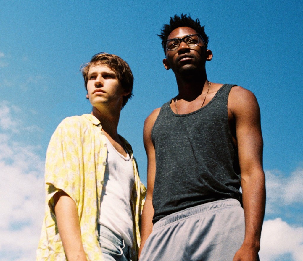 Image is from the film 'Boy Meets Boy' (2021). Two boys stand beside one another, looking out into the horizon. One is wearing a yellow shirt, the other a grey tank top.