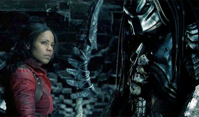 Sanaa Lathan as Alexa Woods in Alien vs. Predator (2004). Alexa is wearing a red jacket and is looking into the distance. Next to her, stands a large, masked figure.