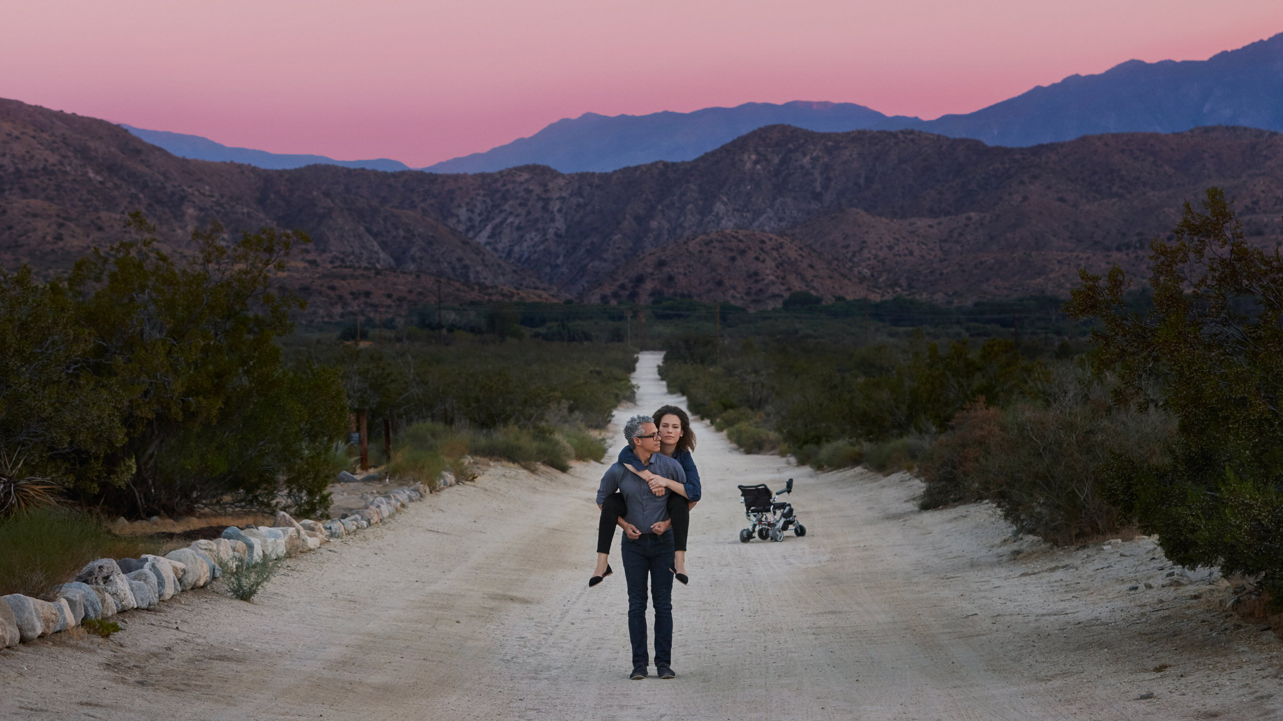 Image is from the documentary 'Unrest' (2017). Against a pink sunset and a rocky landscape, Jennifer Brea is held up by her husband in the middle of a dirt road. Behind them is Jennifer's wheelchair.