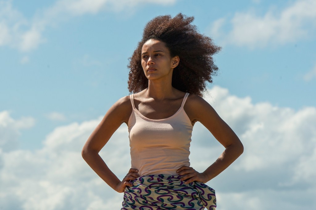 Capitu stands, hands on her hip, with the sky in the background.