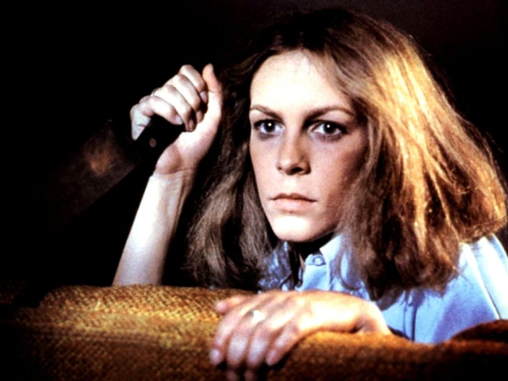 Jamie Lee Curtis as Laurie Strode in Halloween (1978). Laurie is holding a knife and is peering from behind a chair.