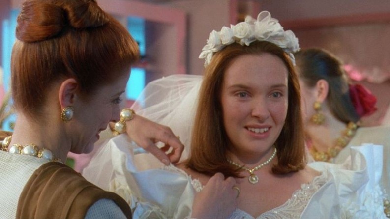 Image is from the film 'Muriel's Wedding' (2004). A young woman smiles as she is fitted into a wedding dress by an older woman.