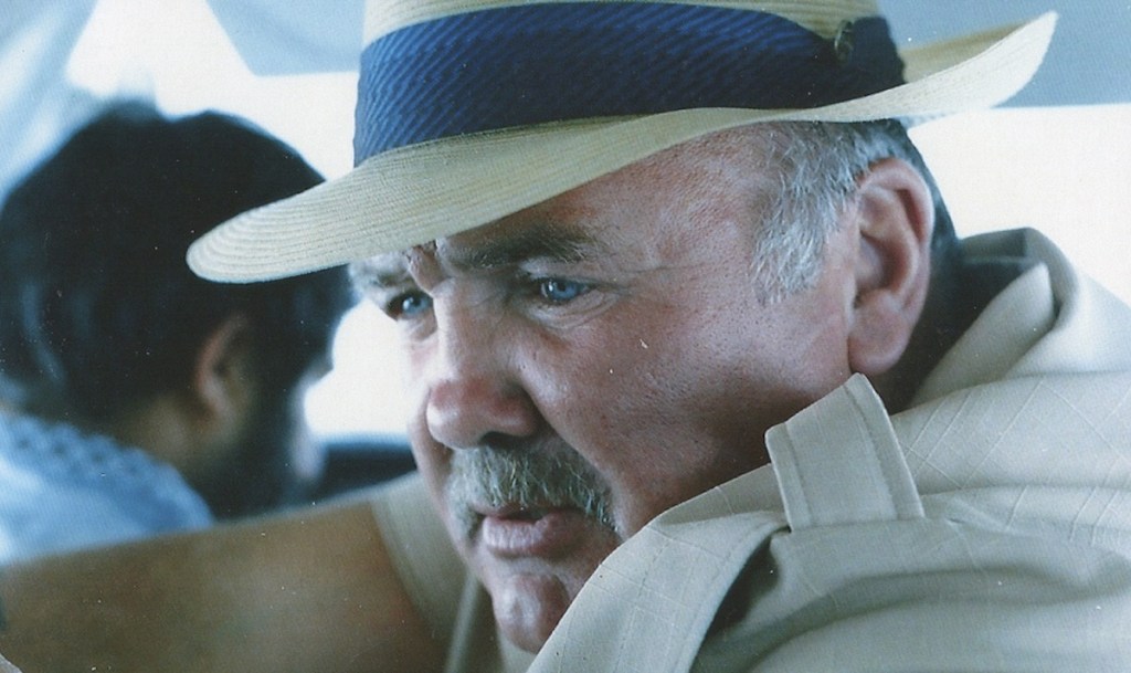 Image is from the film 'Killing Escobar' (2021). A man wearing a beige shirt and a straw hat sits in a car.