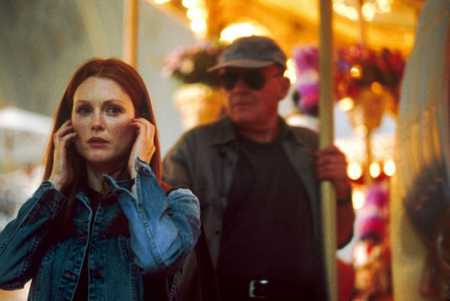 Image is from the film 'Hannibal' (2001). Hannibal sits, masked by shades and a cap, a merry go round behind an oblivious Clarice.