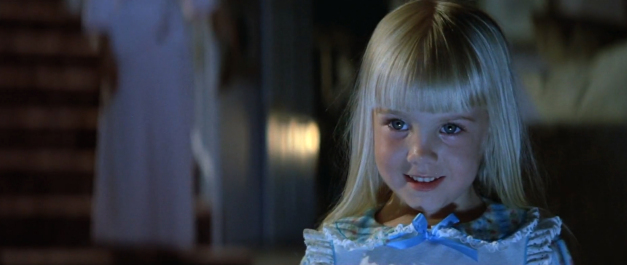 Heather O'Rourke as Carol Anne in Poltergeist. A young child with bright blonde hair and is wearing a blue nightdress smiles in a dimly lit hallway.