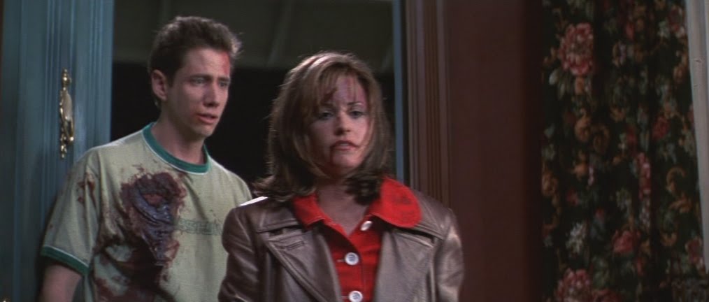 Courteney Cox as Gail Weathers and Jamie Kennedy as Randy Meeks in Scream (1996.) Gail looks down as she walks out of a house, a gash of blood on her lip and forehead. Behind her, Randy - who is also covered in blood - tries to stop her.