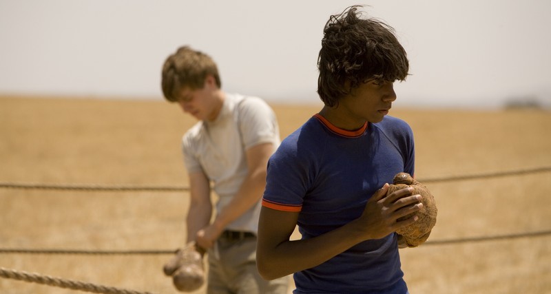 Image from the film September. Ed stands in the back of their makeshift boxing ring, putting on a worn boxing glove. In the front, Paddy holds his hands together - one is gloved whilst the other isn't.