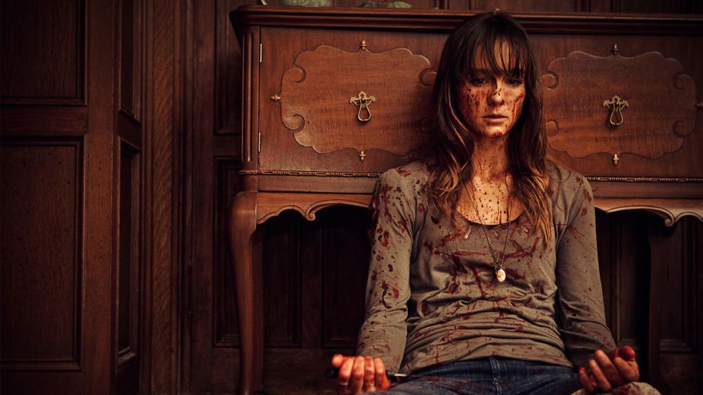Sharni Vinson as Erin in You're Next (2001). Erin sits on the floor, leaning against a cabinet. She looks at her hands, which like the rest of her, are covered in splatters of blood.