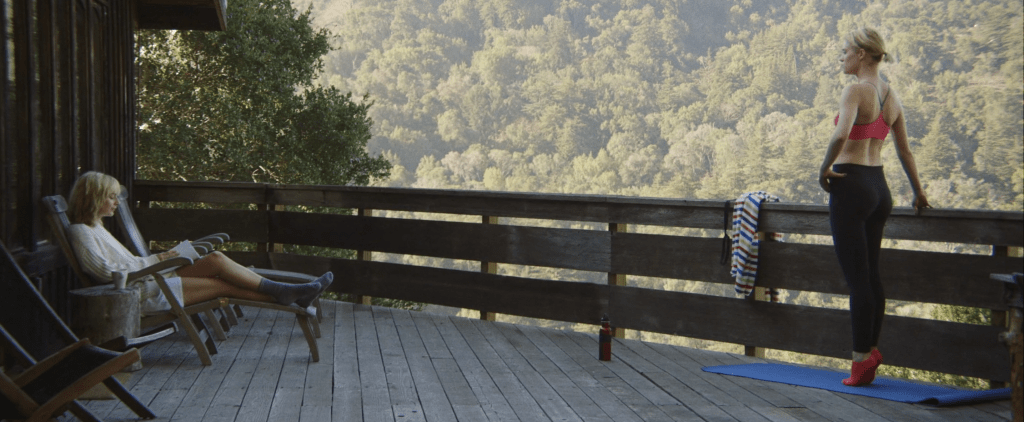 Image is a wide shot with Beth to the far left and Anna to the far right as they’re outside. Behind them is the Big Sur landscape, with thick woods and never-ending trees. Beth is lying with her feet up on a sunbathing chair made of wood as she’s flicking through a new script. Anna is wearing black tights along with socks and a sports bra in a bright pink colour. She’s standing on her toes on a yoga mat, exercising, holding one hand on her hip and the other helping her to balance as she’s holding onto the wooden rail next to her.  